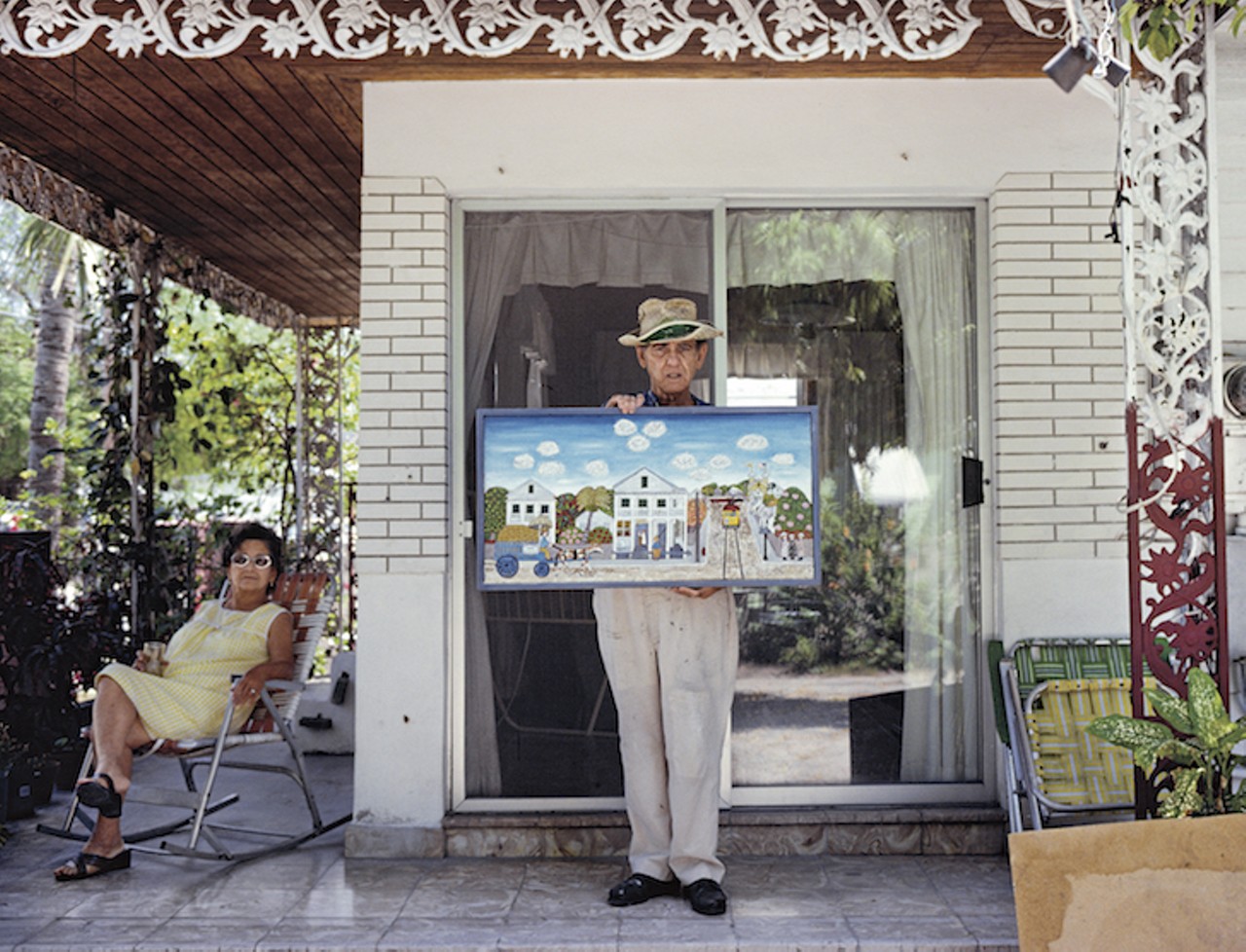 Folk artist proudly displays a recent painting on the veranda of his house in Key West, Florida. The painting depicts an idealized view of old Key West or Havana. Folk Art is also caller Outsider Art. His wife relaxes nearby. The man was born in Cuba. 1981.