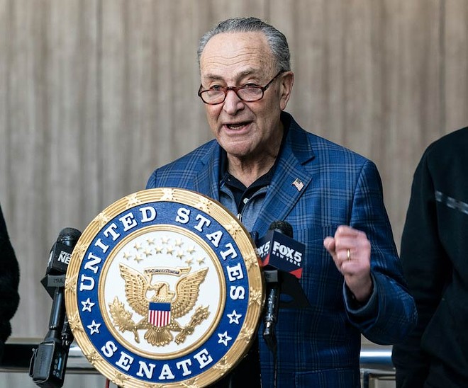 Senator Chuck Schumer stands at a podium with the United States Senate seal. He is wearing a blue plaid sorts coat, reading glasses and a flag lapel pin. On fist is clenched and raised slightly above elbow height.