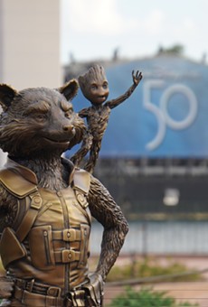 Rocket and Groot Fab 50 statue at Epcot with the 50th-anniversary logo seen on a Harmonious barge LED screen behind them.
